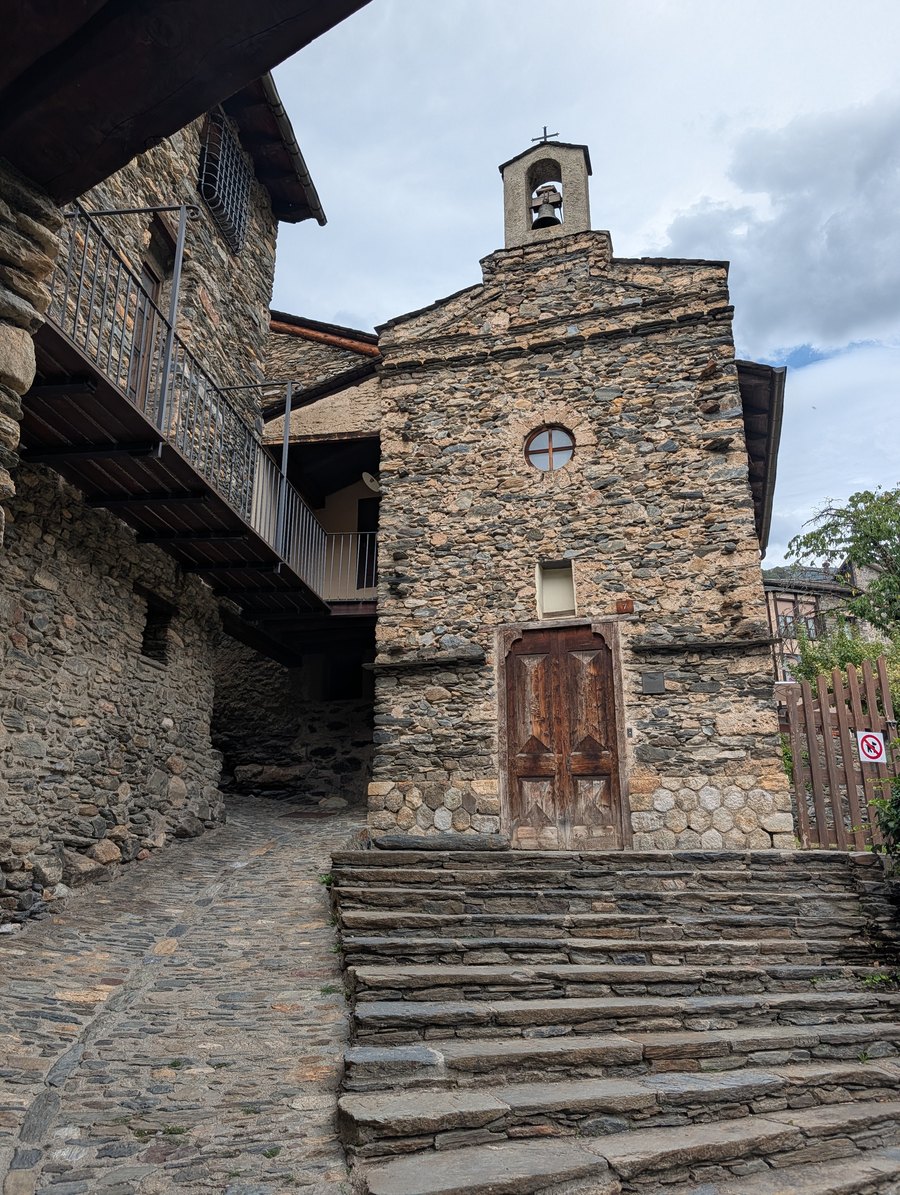 A small village chapel of rough stone with a bell-gable and single bell, a round window above a heavy wooden door, reached by a broad flight of slate steps rising from a cobbled lane between stone houses.