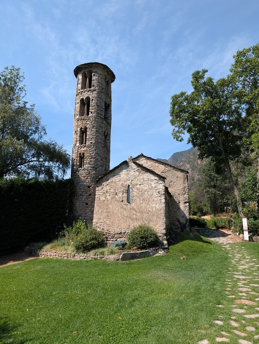 A small stone church with a tall round bell tower of rough masonry, its upper storeys pierced by paired arched windows, standing on a lawn with a cobbled path leading past it and a wooded mountain behind.