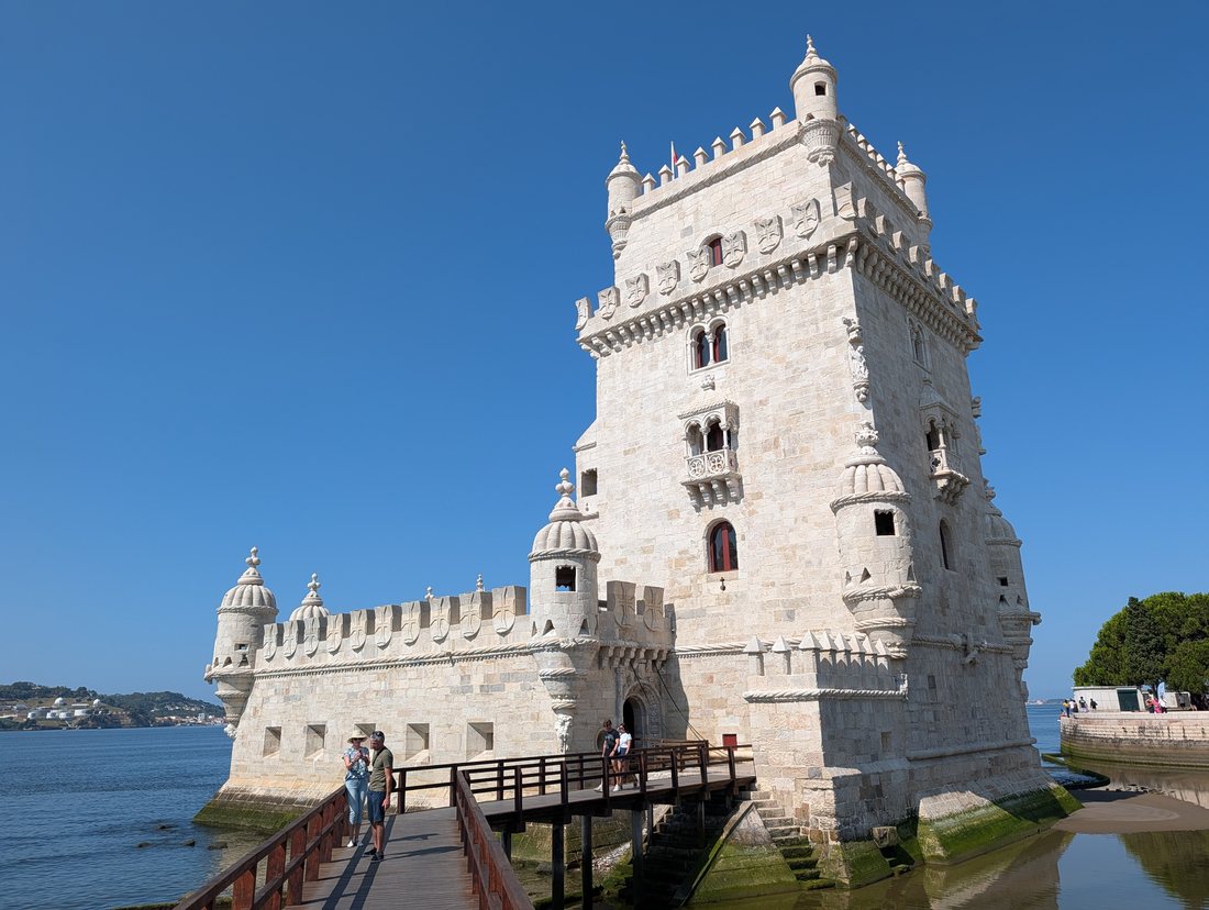 The Torre de Belém photographed from its timber causeway under a clear sky: pale limestone tower with ribbed cupolas on its bartizans, the low hexagonal bastion in front of it, standing in shallow water.