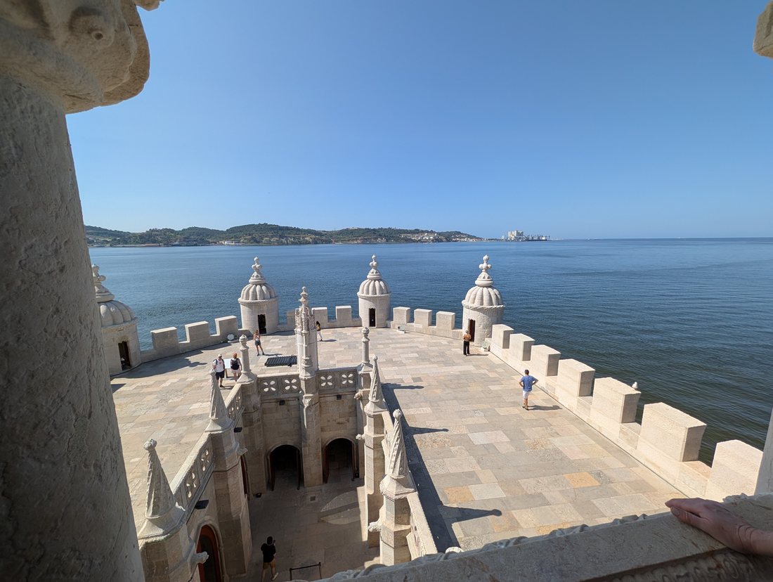 Looking down from the top of the tower onto its terrace: pale stone paving, merlons along the parapet, four ribbed bartizan cupolas, and the wide blue Tagus running out to the far bank.
