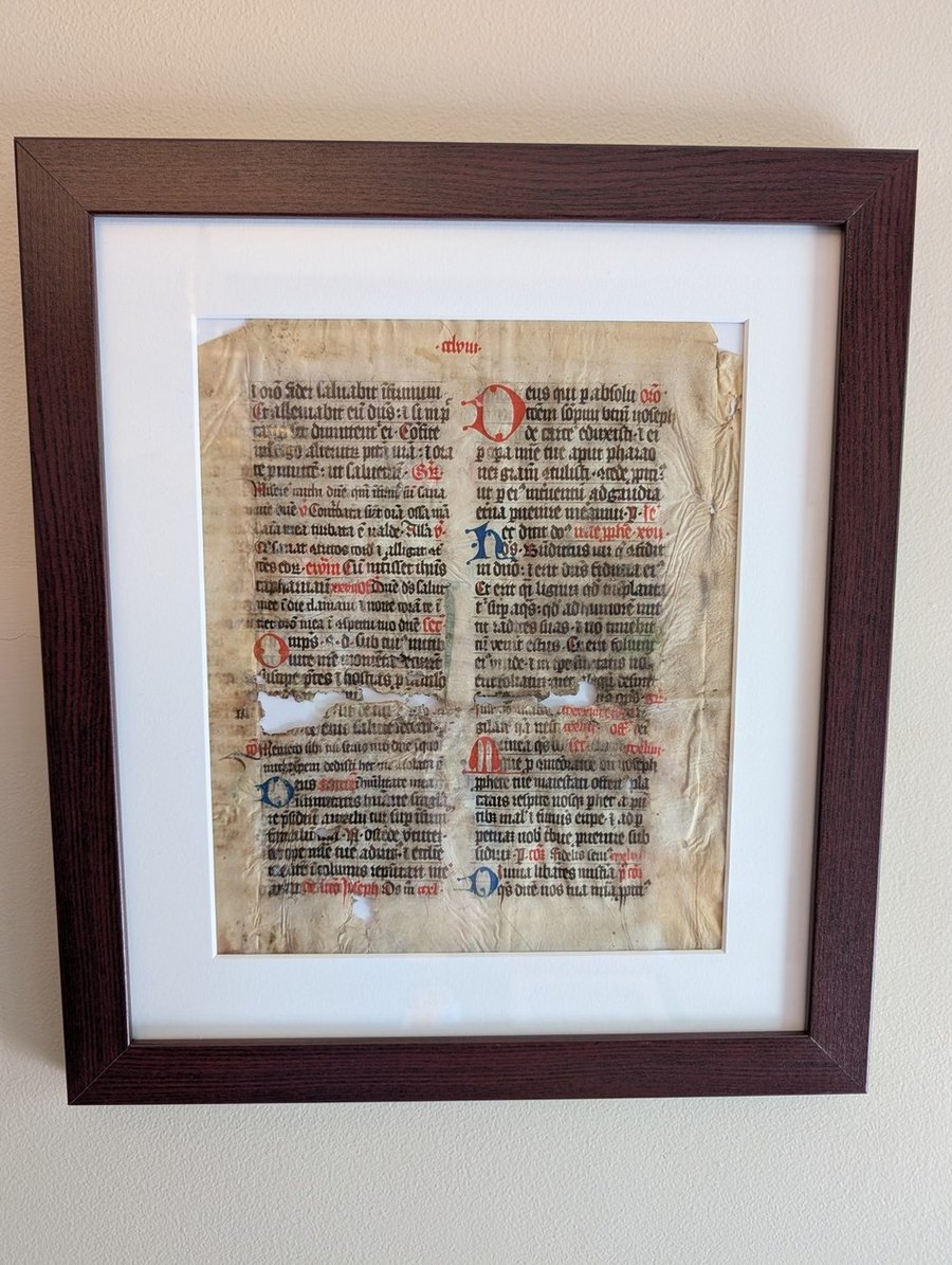 The recto of a medieval vellum leaf, framed behind glass: two columns of gothic script with red and blue initials, and losses torn through the parchment near the centre.
