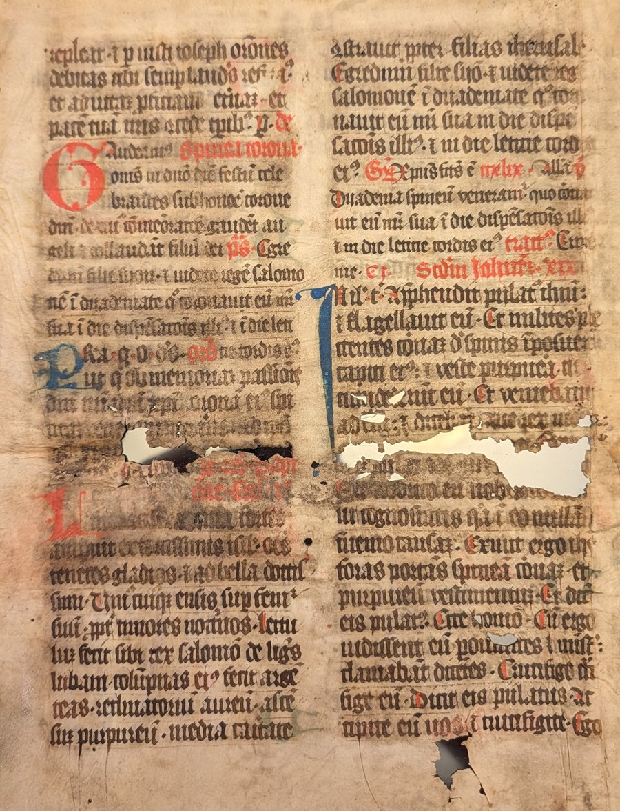 The verso of the same leaf, unframed: two columns of gothic textura on vellum with red rubrics, a large blue initial, and losses through several lines of text.