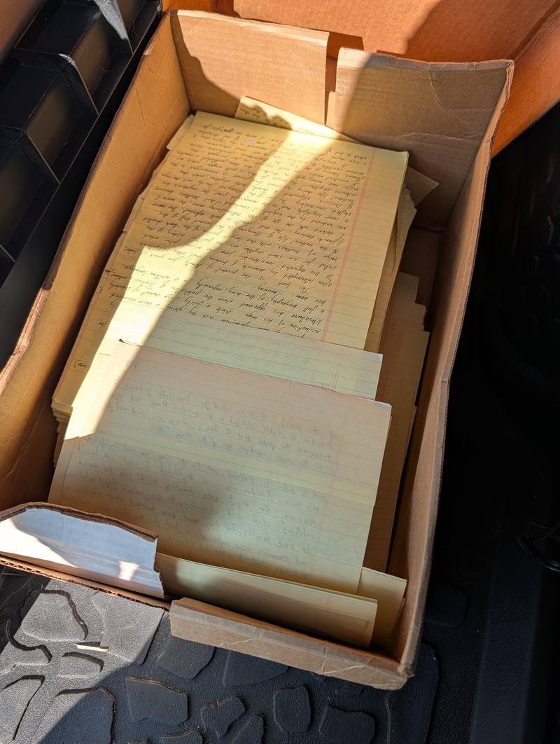 A cardboard box of handwritten manuscript pages on yellow legal paper, photographed in sunlight in the back of a car.