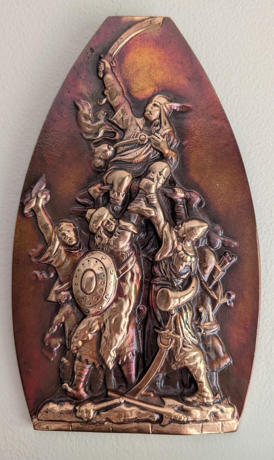 A cast bronze relief plaque in a tall shield shape with a pointed ogee top, hanging on a pale wall. A warrior stands on a shield held up by a group of men below him, his sabre raised overhead; around them a round shield, a drinking horn, a bow and quiver, raised arms, and at the foot a fallen figure among crossed weapons. The raised surfaces are polished bright against a red-brown patina.