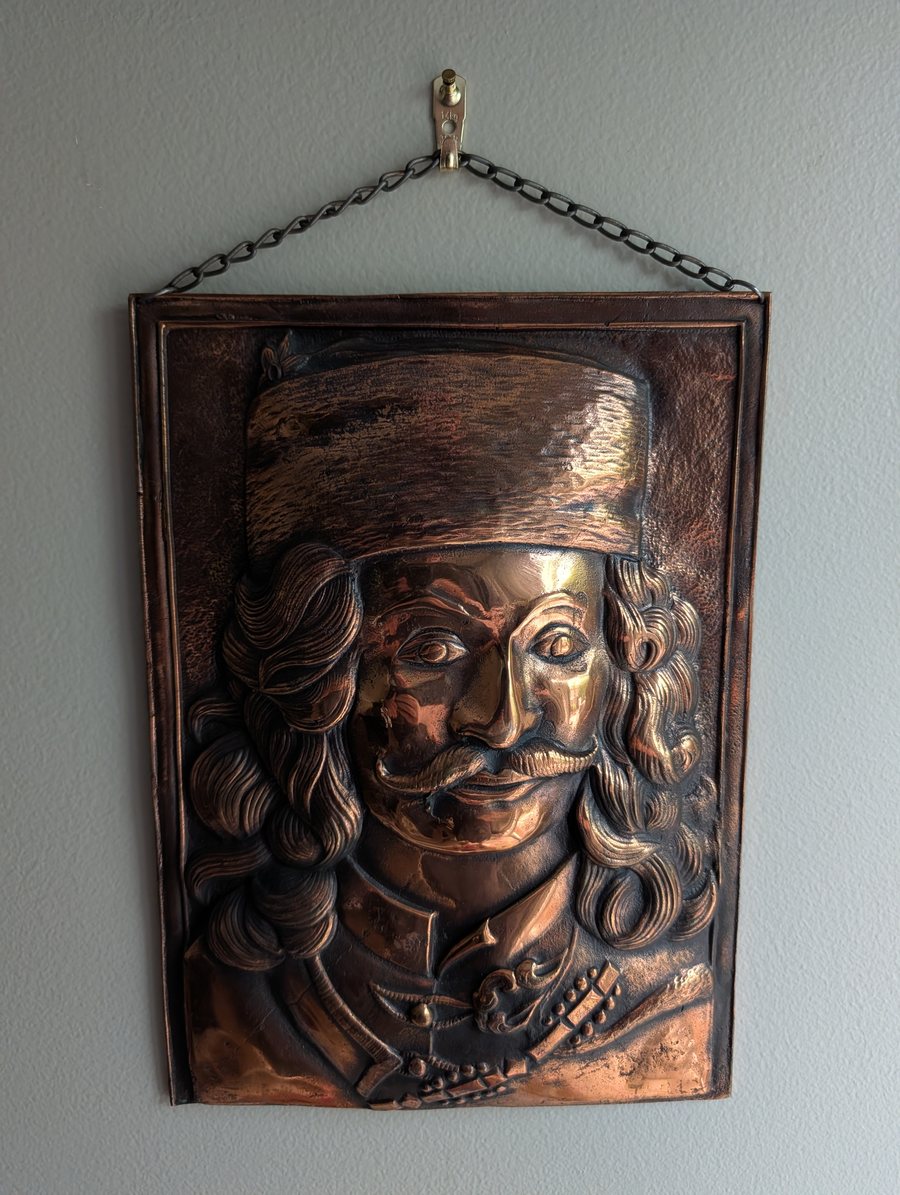 A brass bas-relief plaque hanging on a wall from a chain: a bust-length portrait of a man in a tall fur hat, with shoulder-length curled hair, a curled and twisted mustache, and a frogged coat with a beaded sash. The high points of the relief are burnished bright against a dark patina.