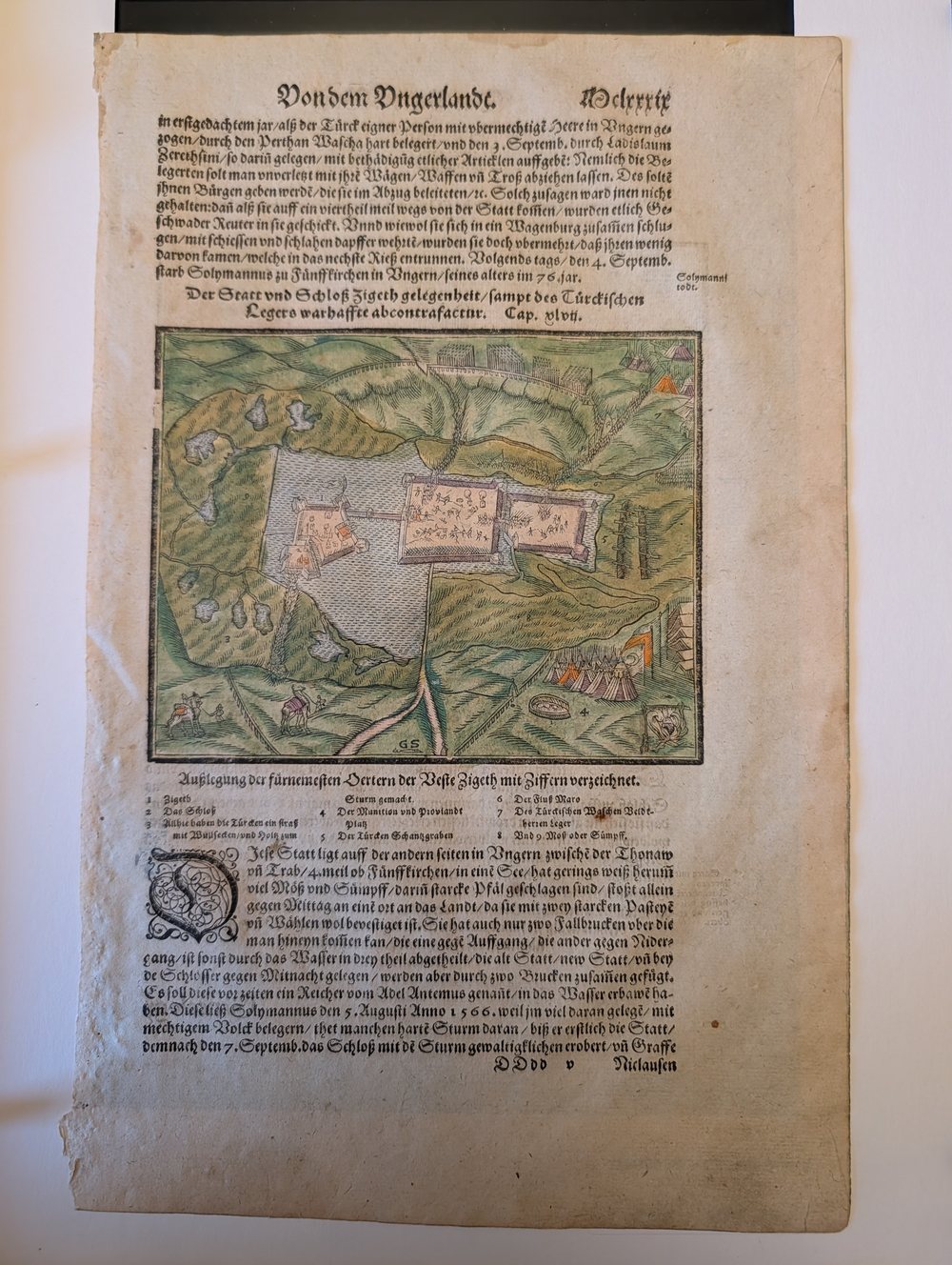 Hand-coloured woodcut view of the fortress of Szigetv&aacute;r surrounded by its lake and marsh, with the Ottoman siege camp and tents at the right, set in a page of German blackletter text.